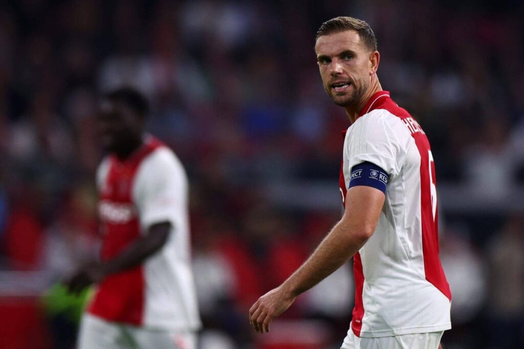 Jordan Henderson, wearing Ajax's white and red home kit with the captain's armband, looks focused during a match as he glances toward the action on the field. A blurred teammate and crowd are visible in the background.