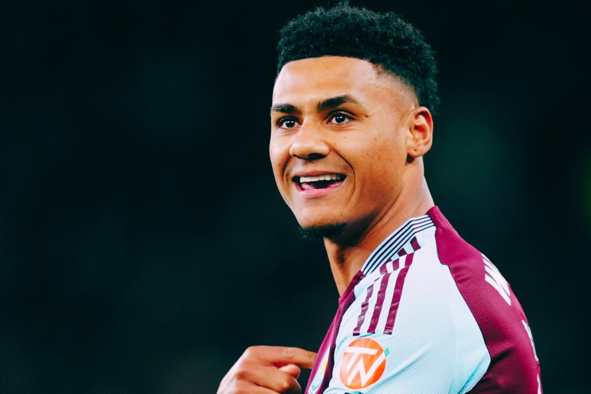 Arsenal interested in Watkins wearing a maroon and light blue jersey, pointing at the logo on his chest. The background is dark and blurred, drawing focus to the player’s cheerful expression.