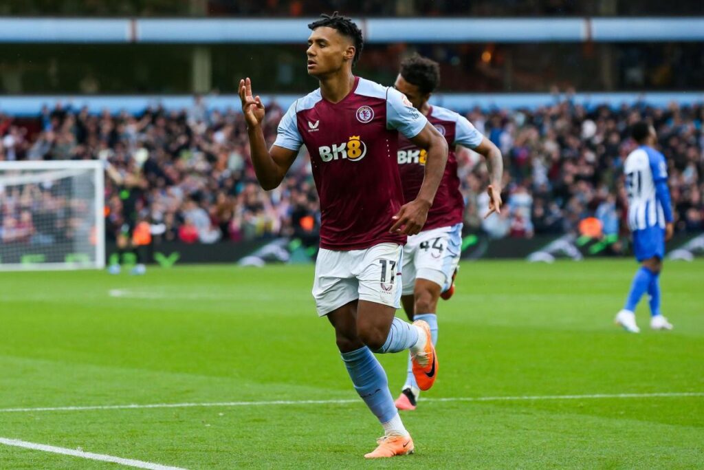 Ollie Watkins celebrates a goal for Aston Villa, wearing the team's maroon and light blue kit. He holds up three fingers as he runs across the pitch, with a vibrant crowd in the background.