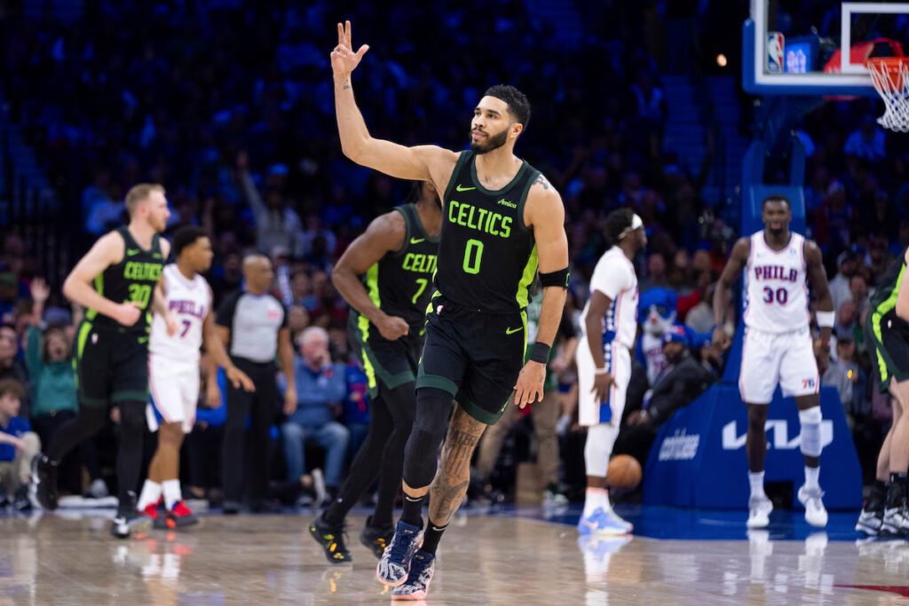 Celtics player wearing a green and black uniform, gestures with three fingers after scoring during an NBA game against the Philadelphia 76ers, with players and fans visible in the background.