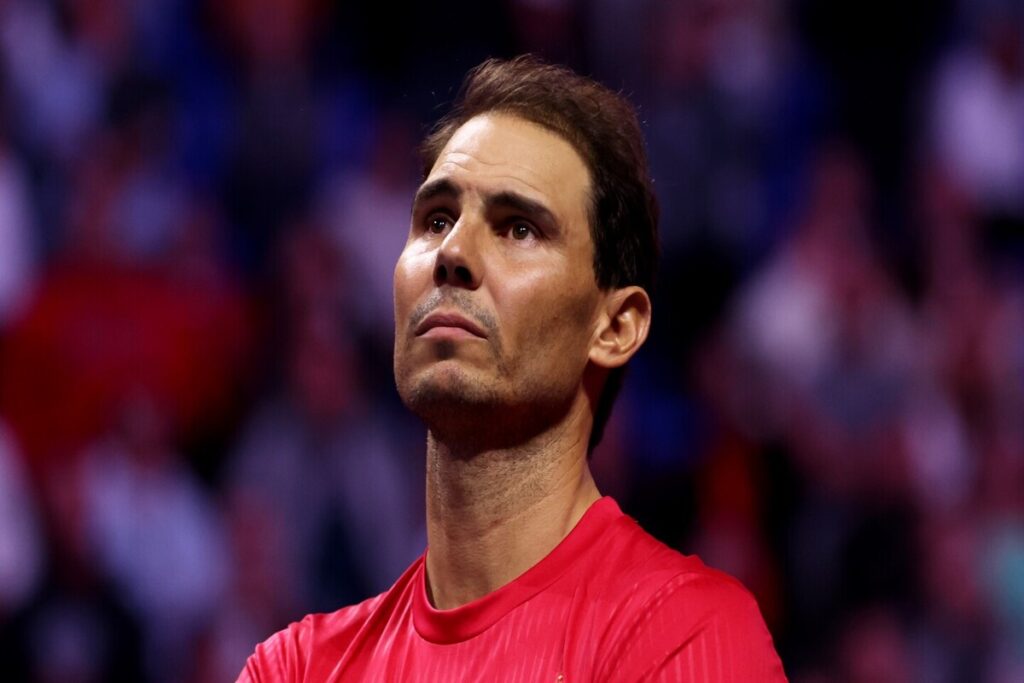 Rafael Nadal, wearing a red shirt looks upward with a contemplative expression, set against a blurred background of an indoor stadium with lights and spectators.