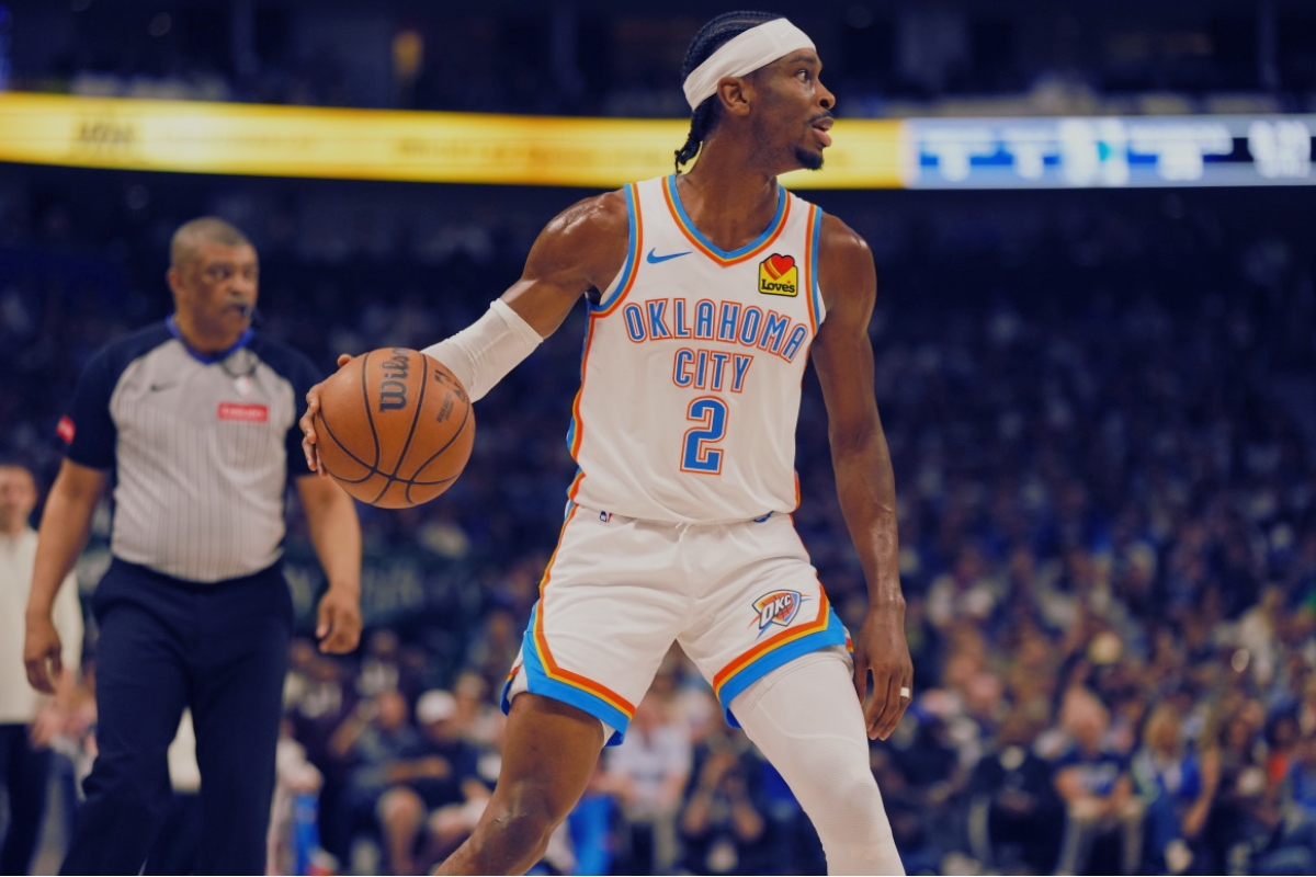 Oklahoma City Thunder guard Shai Gilgeous-Alexander dribbles the ball while scanning the court during a game, with a referee and crowd in the background.